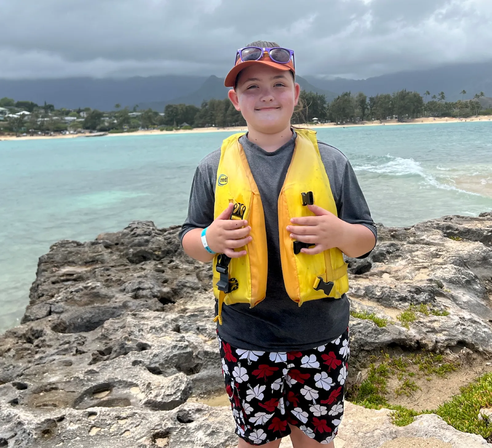 A smiling child in a yellow life vest standing on rocks beside the ocean